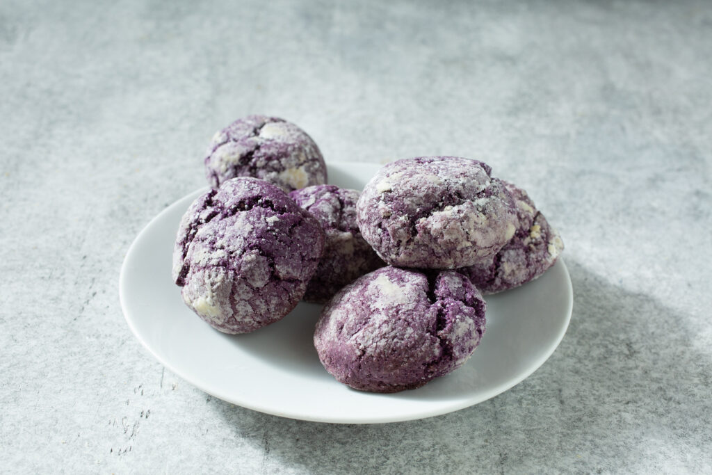 A plate of purple ube crinkle cookies dusted with powdered sugar on a light gray surface. Industrial Ube Puree can be used to make these types of goods.