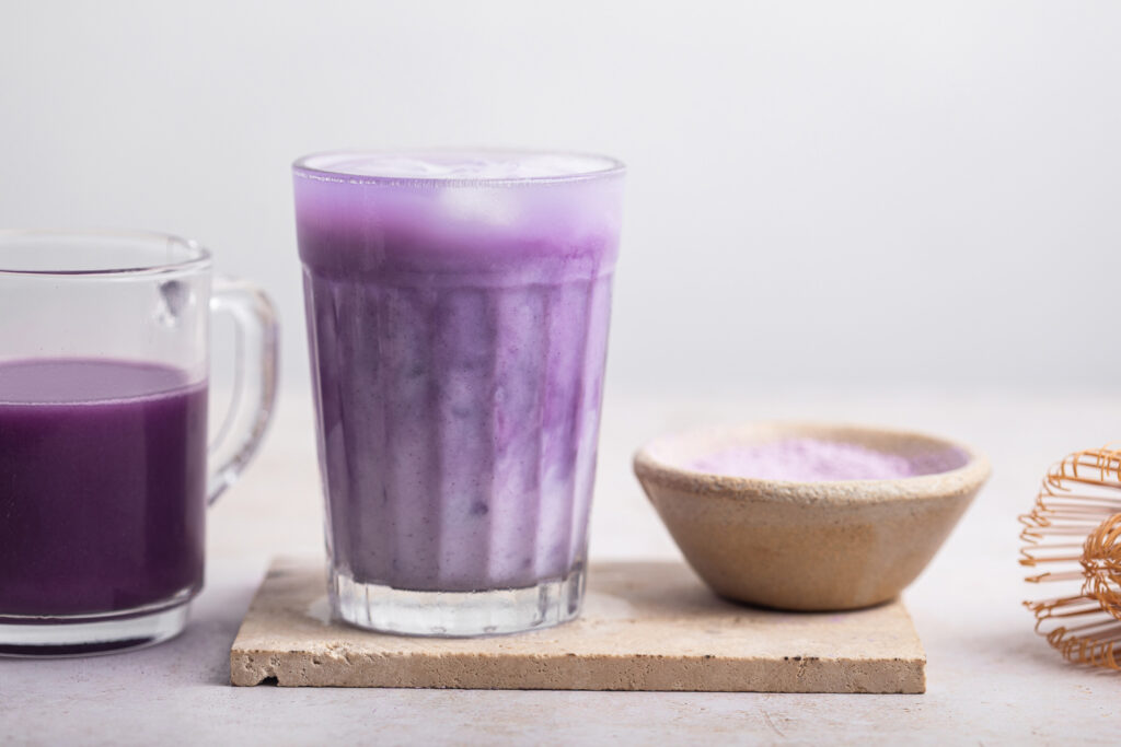 A glass of iced ube latte with layers of purple and white, next to a bowl of ube powder and a whisk.