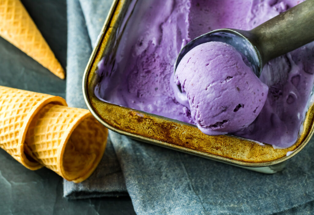 A close-up of creamy purple ube ice cream being scooped from a metal tray with waffle cones beside it.