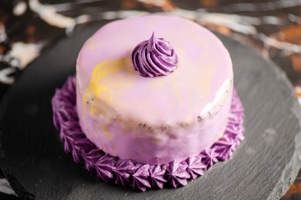 A glossy purple ube cake topped with a swirl of frosting, decorated with piped purple cream on a black slate plate.
