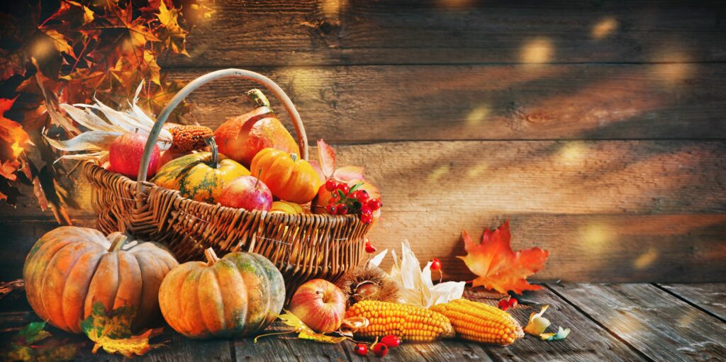 Basket of pumpkins, apples, and corn surrounded by autumn leaves on a rustic wooden table.
