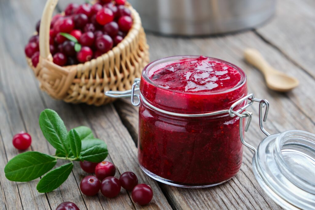 Homemade cranberry jam in a glass jar beside fresh cranberries and green leaves.
